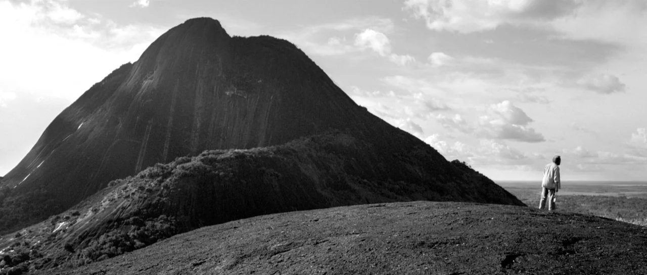 Embrace Of The Serpent (2015) movie still: wide — A stark mountain ridge overlooks a vast jungle under a cloudy sky. An older man stands alone at…