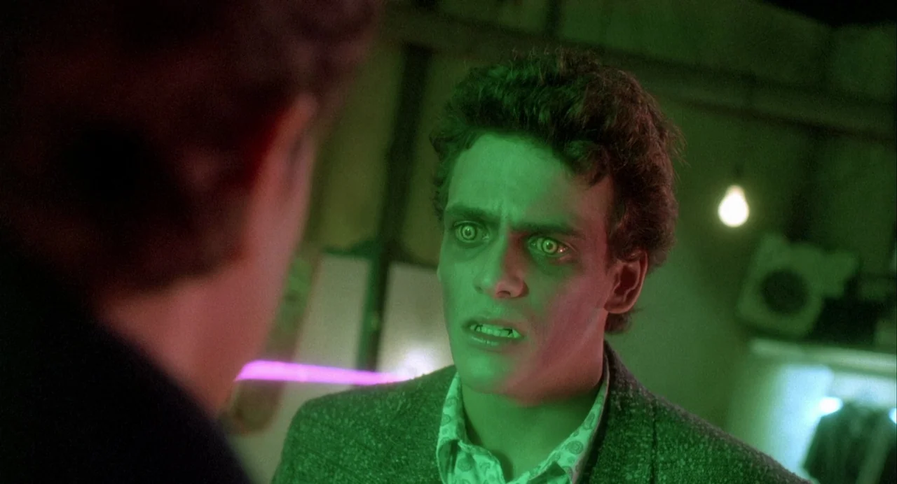 Vamp (1986) movie still: medium — An interior space lit by neon green light with blurred background structures. A young man stares intently at an…