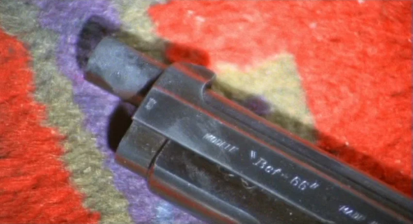 A Lizard In A Woman’s Skin (1971) movie still: closeup — A close-up view of a metal firearm lying on a multi-colored textured textile fabric. The…