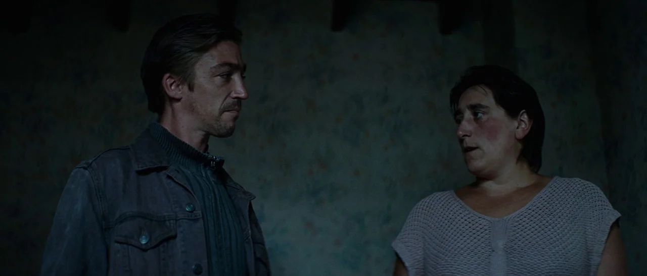 Outside Satan (2011) movie still: medium — A dark indoor space with floral wallpaper is visible behind the two subjects. A man and a woman stand…