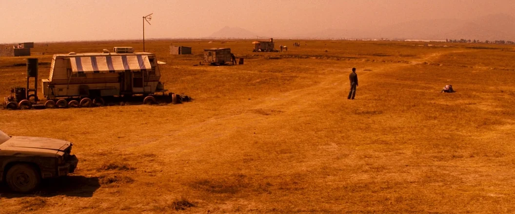 Romeo + Juliet (1996) movie still: wide — A vast, arid, orange-tinted landscape featuring a weathered trailer and an old car. A lone man stands in…