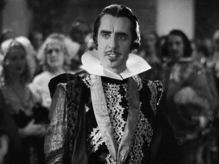 Queen Christina (1933) movie still: medium — A man in a 16th-century style costume stands in a crowded ballroom setting. Other guests in formal…