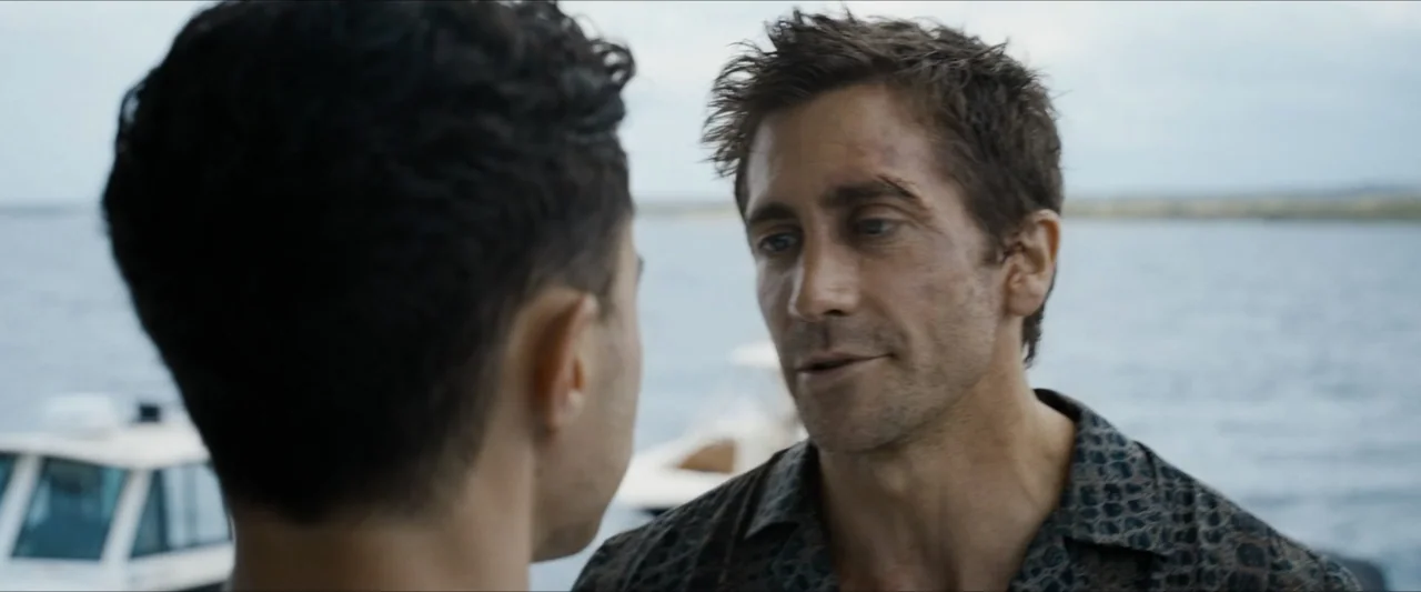 Road House (2024) movie still: medium — A boat deck overlooks a large body of water on a clear overcast day. One man faces another as they engage in…