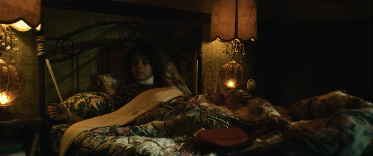 I'll Take Your Dead (2018) movie still: medium — A dimly lit room features a metal bed frame with vintage floral bedding. A woman with dark hair…