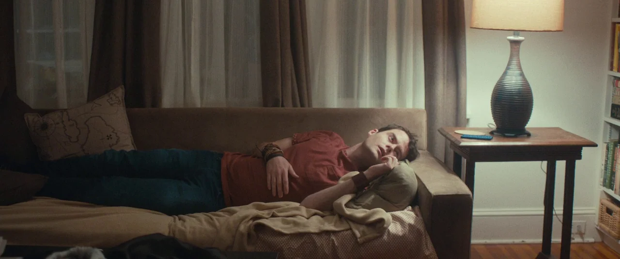 The Skeleton Twins (2014) movie still: medium — A man lies on a brown couch in a room with white walls. He rests on a pillow while looking toward…