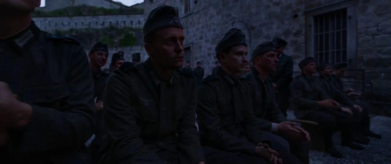 A Hidden Life (2019) movie still: medium — Multiple men in 1940s German military uniforms sit on wooden benches in a stone courtyard. The soldiers…