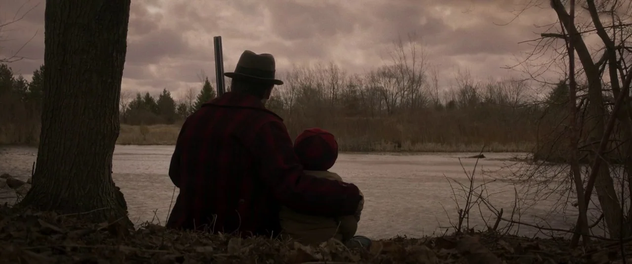 Falling (2020) movie still: medium — A man and a young child sit together on a dry riverbank. The man keeps an arm around the child as they look…