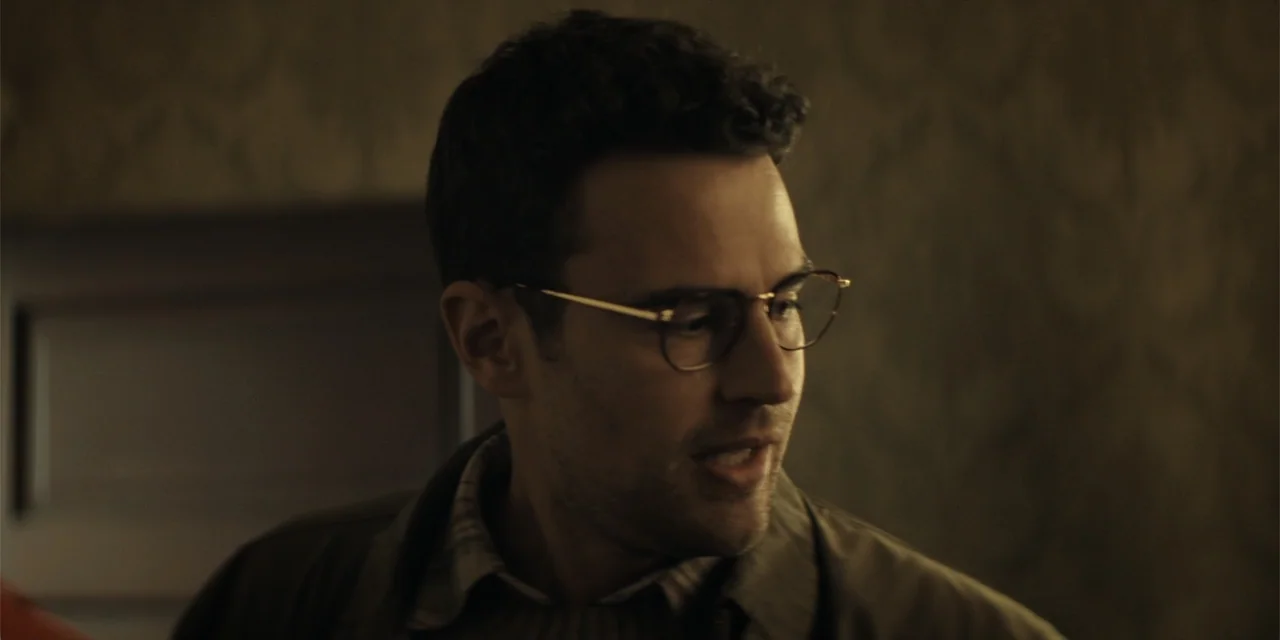 The Monkey (2025) movie still: closeup — An interior space features dimly lit walls with a muted color palette. A man with curly hair and…