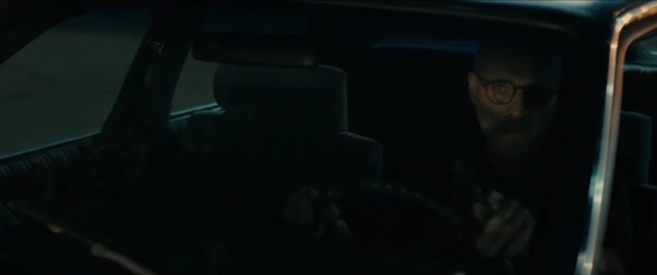 Guns Up (2025) movie still: medium — A dark vehicle interior features a single driver in the night. The driver holds the steering wheel while…