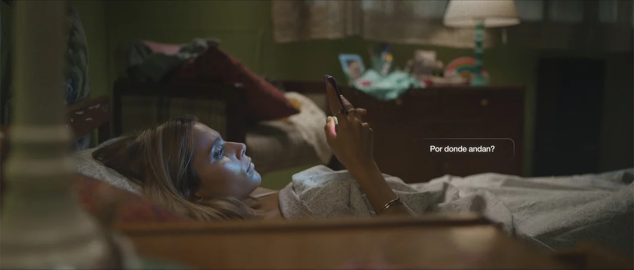Nahir (2024) movie still: medium — A dimly lit bedroom features green walls and a wooden bed frame at night. A young woman lies in bed while holding…