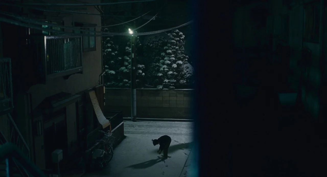 Shoplifters (2018) movie still: wide — A narrow residential street at night is covered in a layer of fresh snow. A lone person stands in the middle…