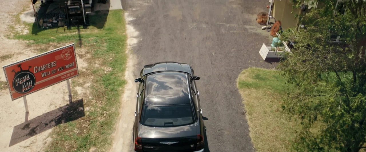 Great White (1998) movie still: wide — An overhead view of a black car parked on a dirt lot beside a billboard. A rectangular sign for an air…