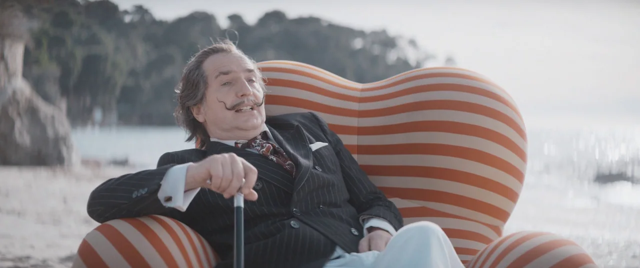 Daaaaaalí! (2023) movie still: medium — A man sits on a large striped sofa placed directly on a sand beach. The background features a blurred…