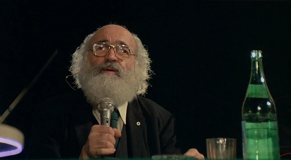 The Voice of the Moon (1990) movie still: medium — An elderly man with a long white beard speaks into a microphone at night. A glass water bottle…