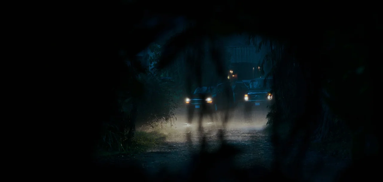 Triple Frontier (2019) movie still: wide — A dark, rain-drenched dirt road is framed by silhouetted foliage in the foreground. Two vehicles with…