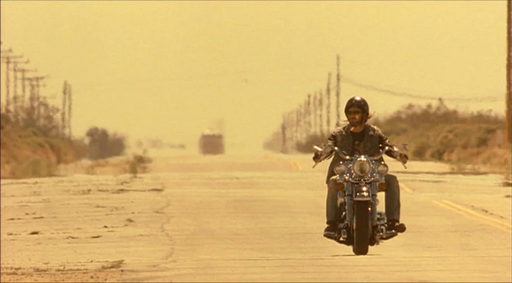 Erin Brockovich (2000) movie still: wide — An endless stretch of cracked asphalt runs through a dry, barren desert landscape. A lone motorcyclist…