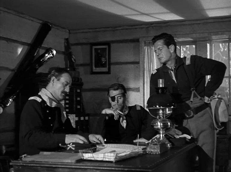 Rio Grande (1950) movie still: medium — A rustic military office features log walls and a large desk piled with documents. Three soldiers in dark…