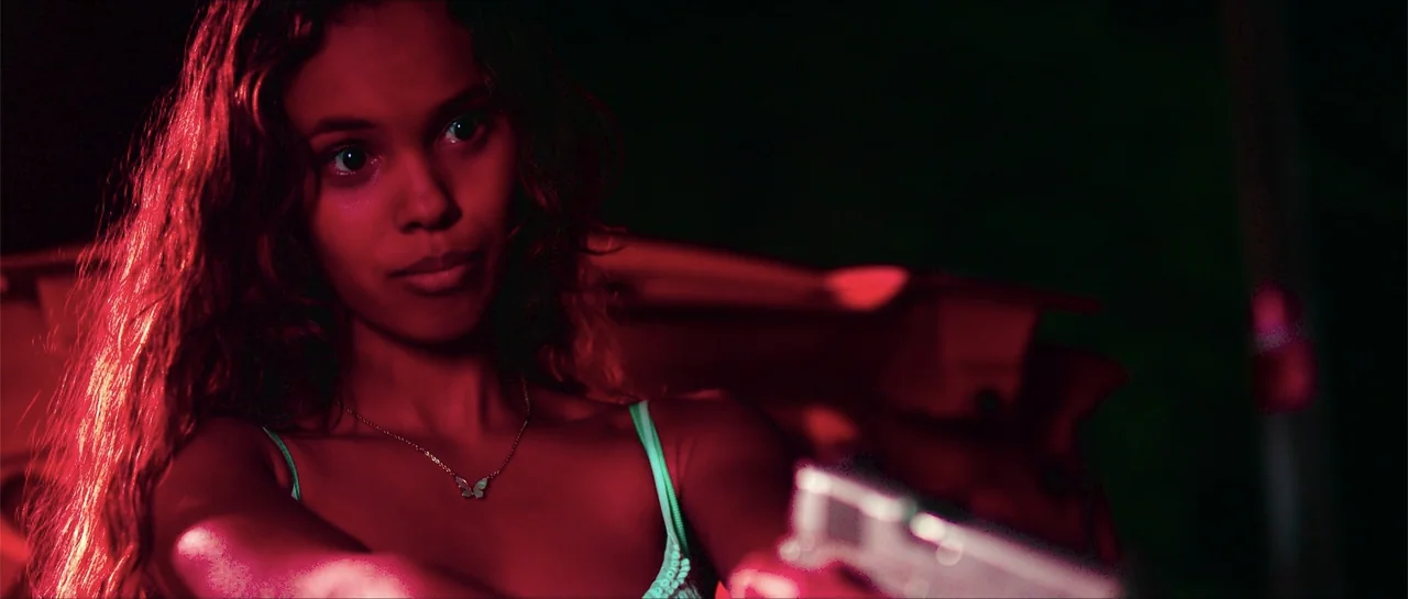 68 Kill (2017) movie still: medium — A dark vehicle interior is bathed in a harsh red light source. A young woman with long hair aims a silver…