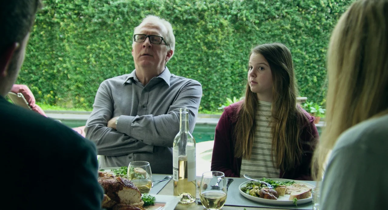 Deep Water (2022) movie still: medium — A patio setting features a dining table with plates of food and wine. An elderly man and a young girl sit at…