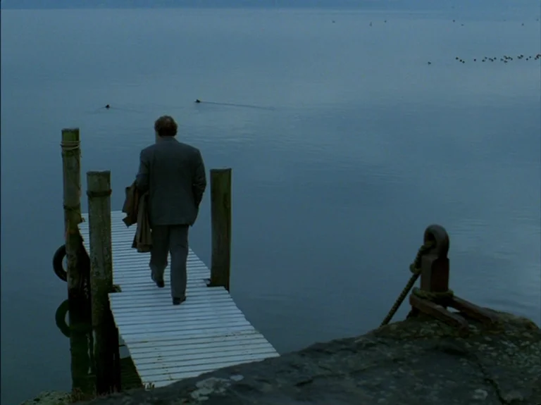 Helas Pour Moi (1993) movie still: fullbody — A wooden pier extends into a vast, calm body of water at dusk. A middle-aged man in a suit walks away…