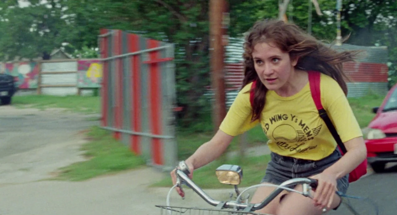 Mr. Roosevelt (2017) movie still: medium — A suburban street features a corrugated metal fence with red and grey panels. A young woman cycles toward…