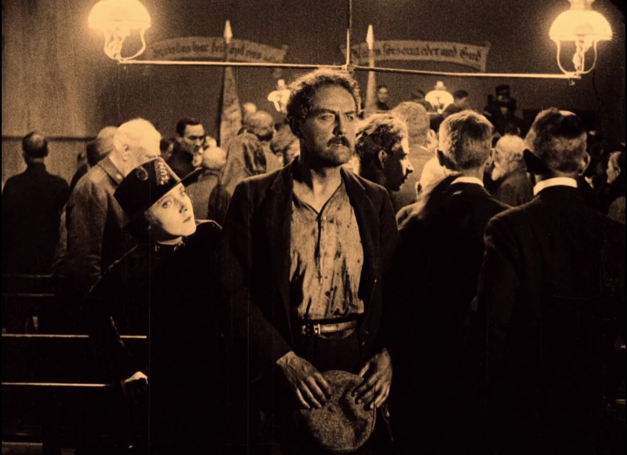 The Phantom Carriage (1921) movie still: medium — A dark hall filled with people is lit by two hanging oil-style lamps. A man and woman stand in the…