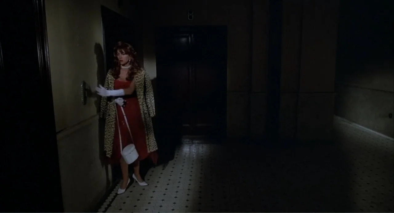 I, Madman (1989) movie still: fullbody — A dimly lit residential corridor with tiled floors and dark wooden doors. A woman stands by a door pushing…