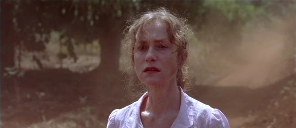 White Material (2009) movie still: medium — A woman stands on a dirt path surrounded by trees and hazy dust clouds. She looks forward while standing…