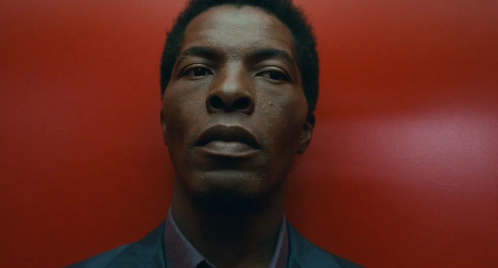 The Limits of Control (2009) movie still: closeup — A man stands before a flat, solid red wall with a matte texture. He faces the camera directly…