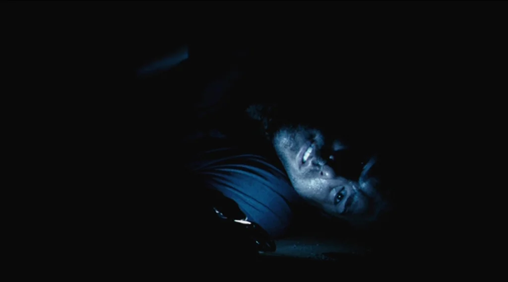 Buried (2010) movie still: closeup — The frame shows a dark, confined space with minimal environmental detail visible. A man lies on his side,…