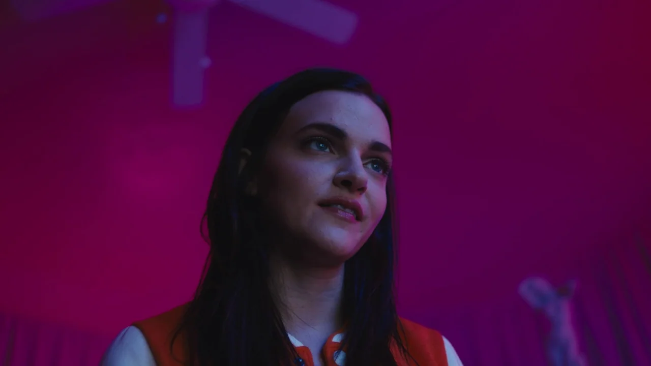 Cam (2018) movie still: closeup — A dimly lit indoor space is saturated with intense magenta light. A young woman with dark hair looks upwards while…