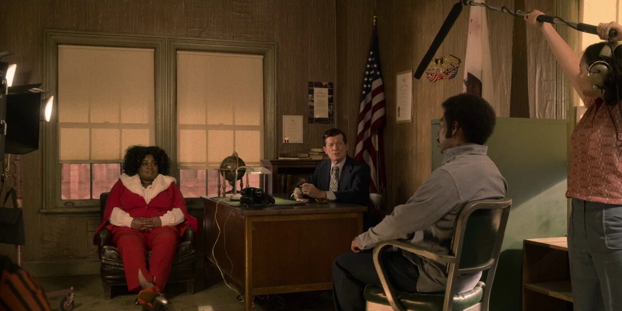 Dolemite Is My Name movie still: wide — An office from the 1970s features wood-paneled walls, an American flag, and period furniture. Three…
