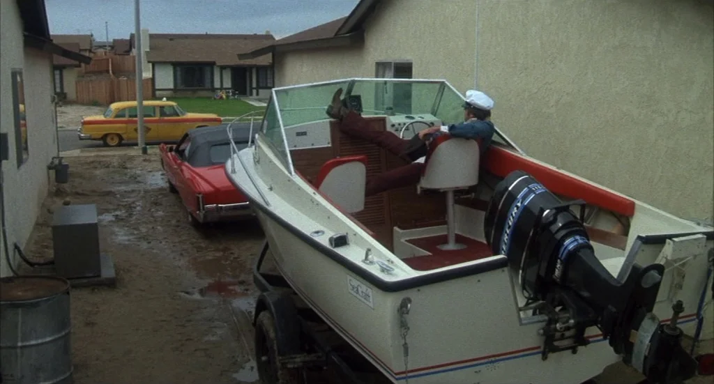 Melvin & Howard (1980) movie still: wide — A suburban residential driveway features a parked boat on a trailer next to houses. A man wearing a…