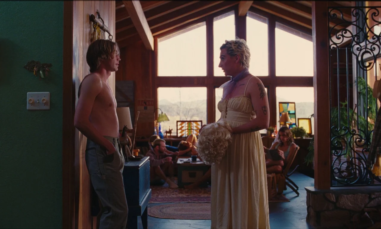 National Anthem (2023) movie still: fullbody — A cabin interior features wooden walls and large windows overlooking a bright exterior landscape. Two…