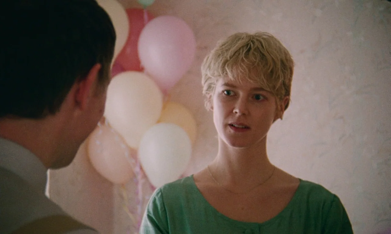 Blue Jean (2022) movie still: medium — A room with textured wall patterns is decorated with a cluster of colorful balloons. A young woman with short…