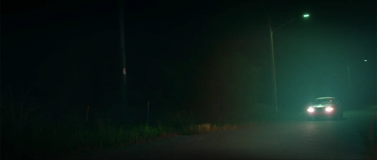 68 Kill (2017) movie still: wide — A lonely paved road stretches into darkness under the dim glow of streetlamps. A car approaches from the distance…