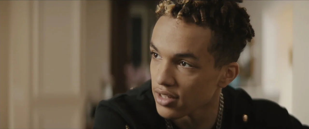 Stalker (2020) movie still: closeup — An interior space with a blurred, light-colored background. A young man with curly hair looks off-camera while…