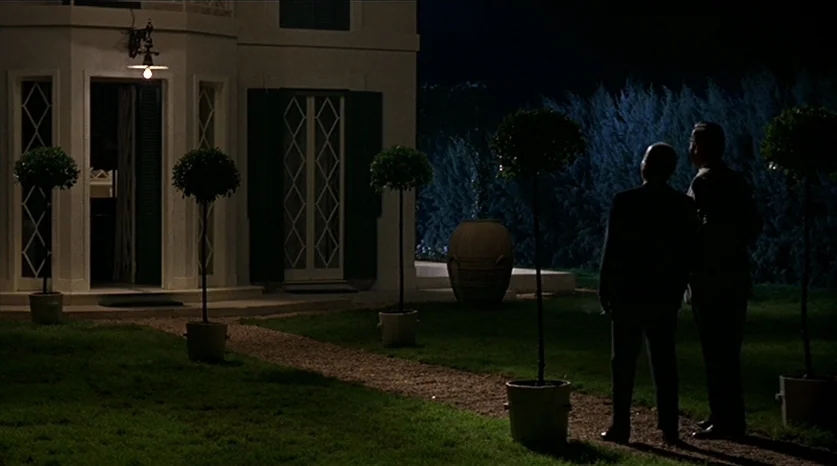 Juliet of the Spirits (1965) movie still: wide — A dark night scene shows a white mansion exterior with a glowing doorway entrance. Two men in suits…