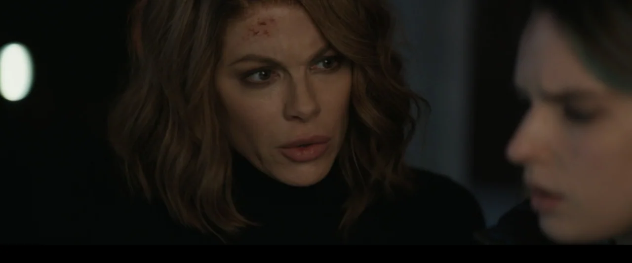 Canary Black (2024) movie still: closeup — A dimly lit indoor space with shadows obscuring most architectural details. A woman with an injured…