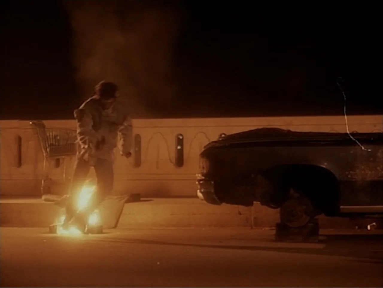 Firepower (1979) movie still: medium — An asphalt surface at night features a flickering fire and a discarded car chassis. A man leaps over the…