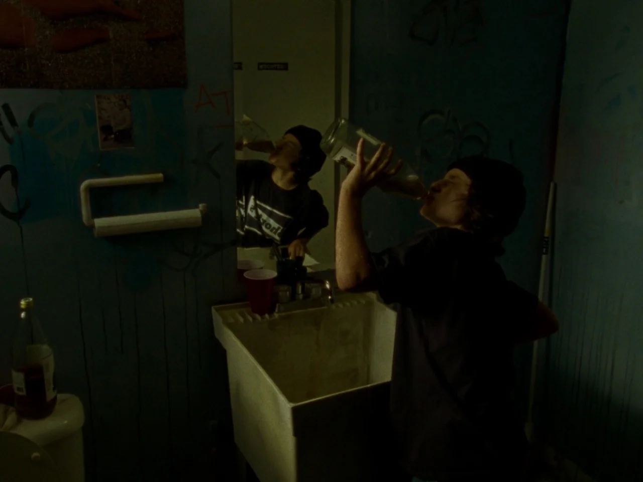 Mid90s (2018) movie still: medium — A dimly lit room features walls covered in dark graffiti and a utility basin. A teenager tilts their head back…