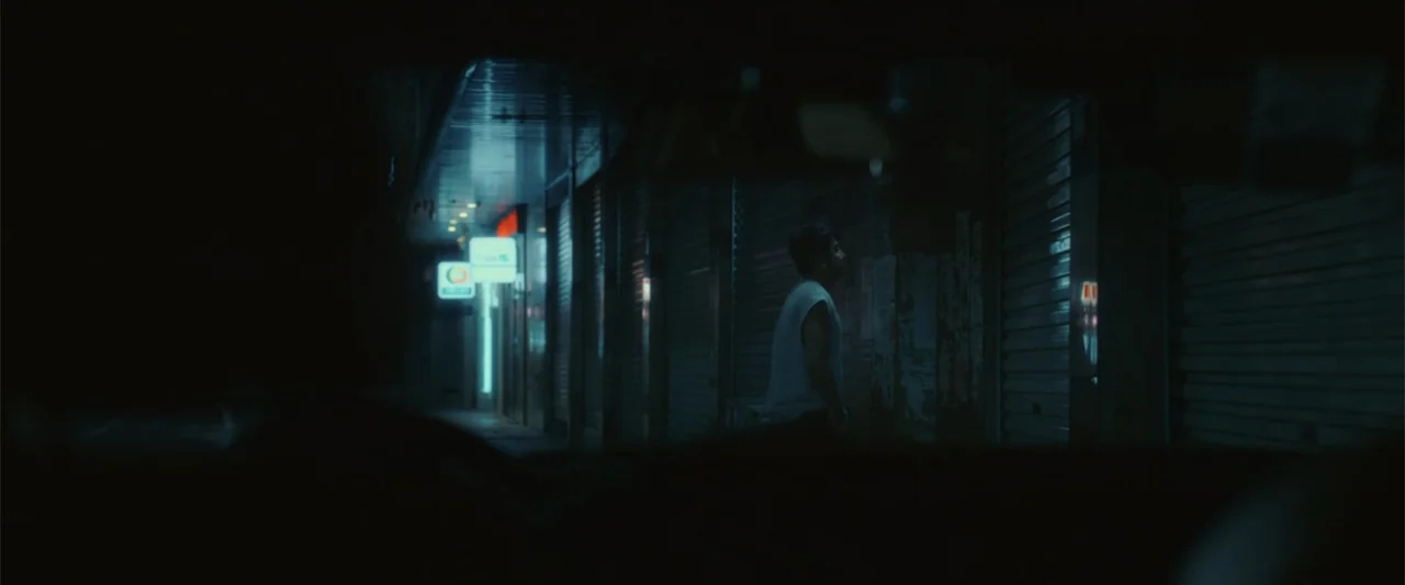 Yolo (2024) movie still: medium — A dark city street at night features a row of closed metal storefront shutters. A man in a sleeveless shirt stands…