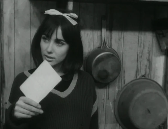 Les Carabiniers (1963) movie still: medium — A dark wooden wall backdrop features two iron skillets hanging on hooks. A woman stands in front of the…