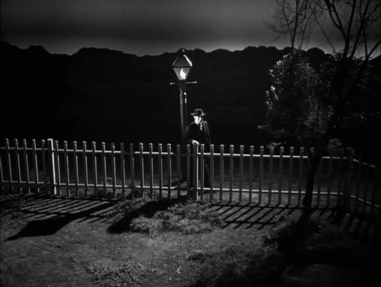 The Night Of The Hunter (1955) movie still: wide — A night landscape features a lone streetlamp positioned on a dirt path. A man in a dark suit…