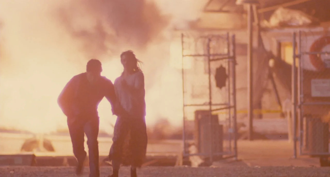 The Big Easy (1986) movie still: fullbody — A fenced-in industrial area is lit by a large, bright explosion in the background. A man and a woman run…