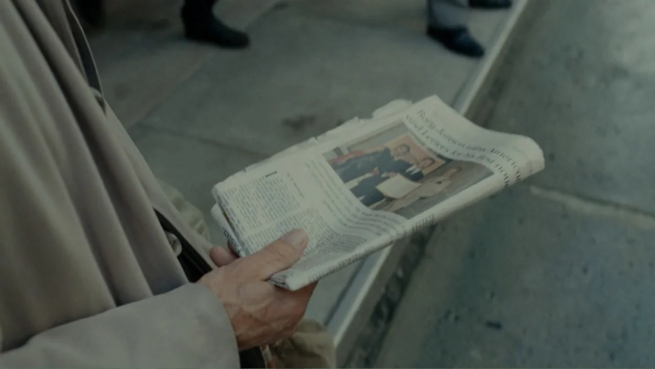 The Words (2012) movie still: closeup — A gray concrete sidewalk is visible from a high angle perspective. A pair of hands holds an open newspaper…