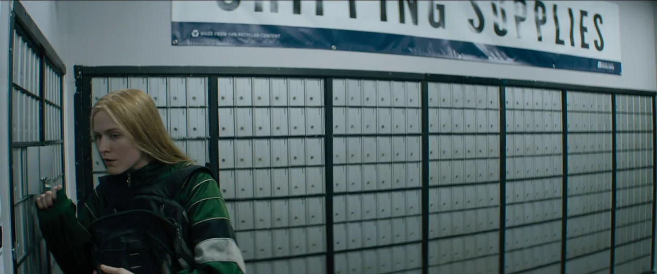 Kajillionaire (2020) movie still: medium — A utilitarian mailroom features rows of uniform grey metal lockers along a plain wall. A young woman with…