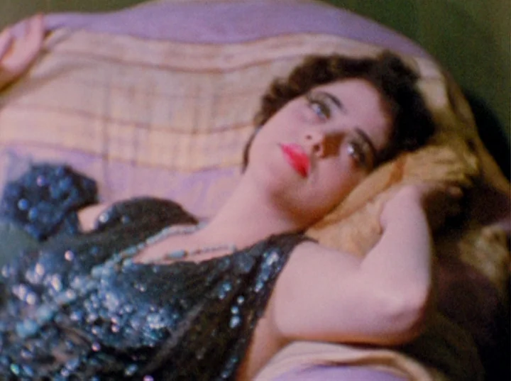 Puce Moment (1949) movie still: closeup — A woman rests on a plush cushioned surface with fabric draped in the background. She lies back on the…
