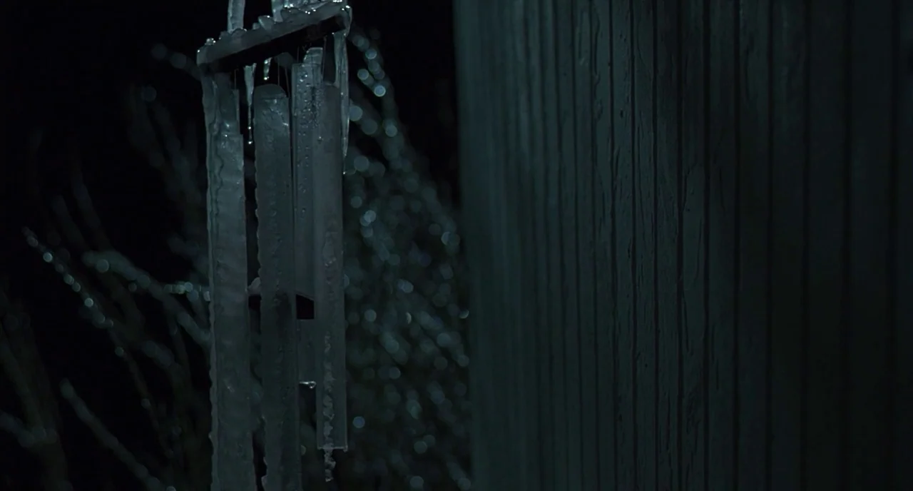 The Ice Storm (1997) movie still: closeup — A set of icicle-covered wind chimes hangs against a dark vertical wooden wall. Blurred tree branches are…