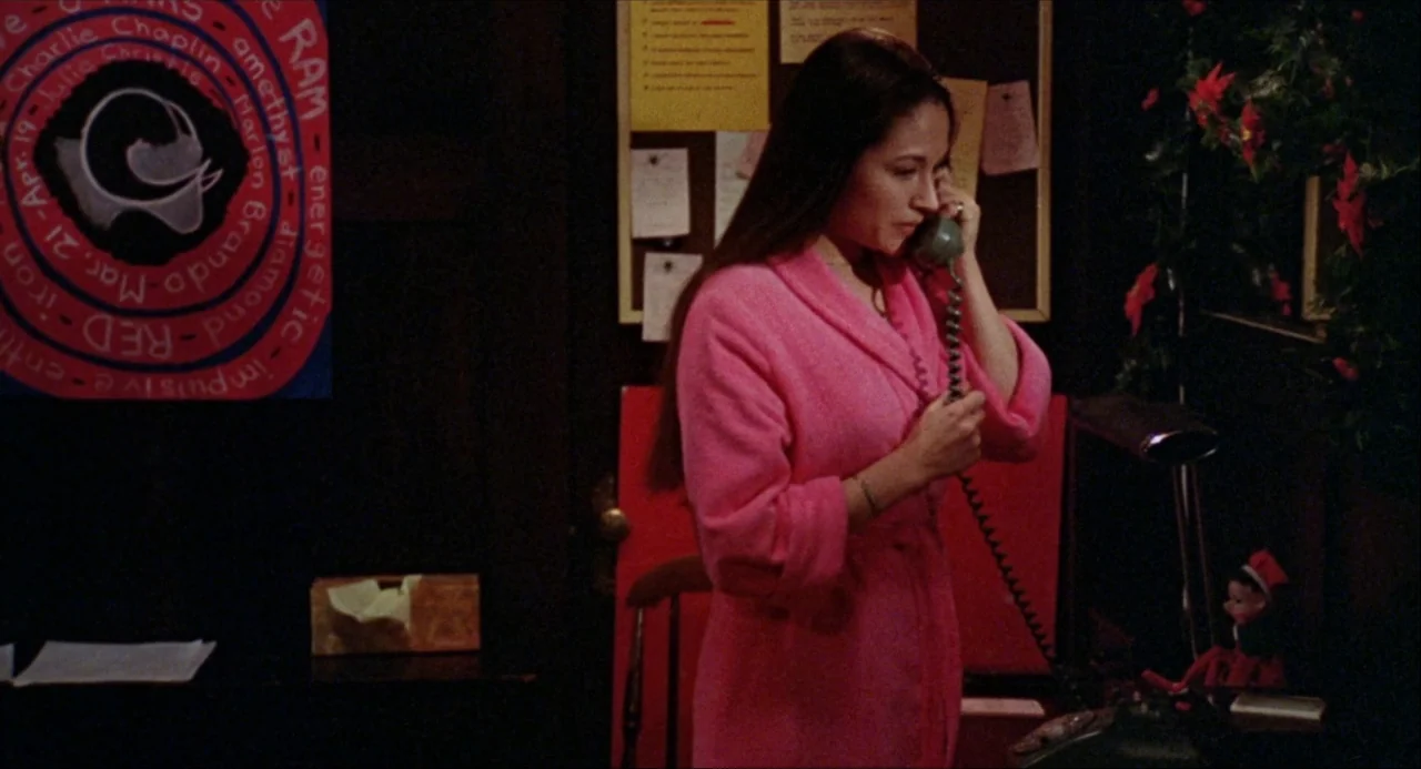 Black Christmas (1974) movie still: medium — A dimly lit room featuring a bulletin board and a red circular wall poster. A woman stands holding a…